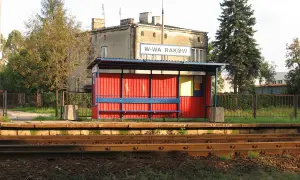 Przystanek kolejowy Warszawa Raków. Czerwona wiata z niebieską ławką i tablicą informacyjną. W tle budynek i wysokie drzewa.