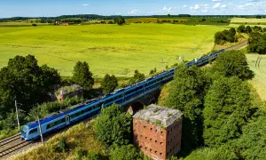 Szynobus Olsztyn Szymany mija ruiny fortu na tle zielonych pól i błękitnego nieba.