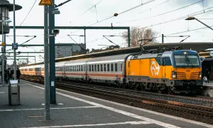 Pociąg z żółtą lokomotywą stoi na stacji, pasażerowie czekają, być może z powodu opóźnienia.