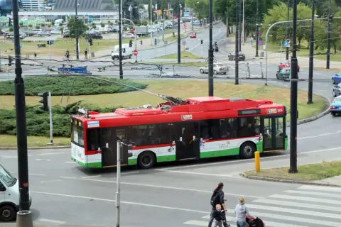 Czerwony trolejbus z napisem "Lublin" przejeżdża przez skrzyżowanie. W tle widać ruch uliczny i budynki.