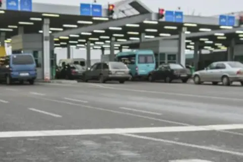 Kolejka samochodów na granicy polsko rosyjskiej. Widać budynki przejścia granicznego, sygnalizację świetlną i różne pojazdy.