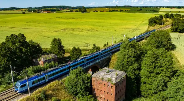 Szynobus Olsztyn Szymany mija ruiny fortu na tle zielonych pól i błękitnego nieba.
