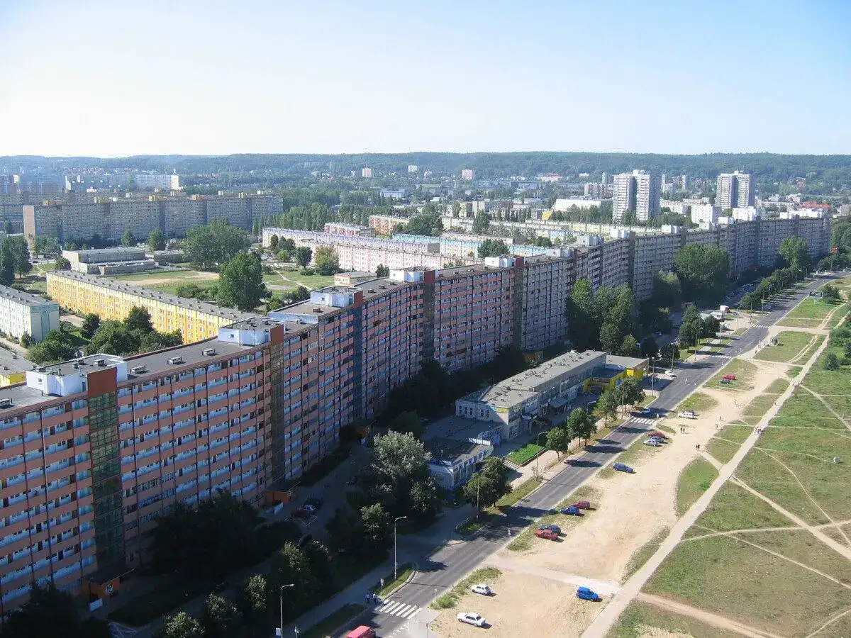 Długi, falisty budynek mieszkalny, znany jako Falowiec Gdańsk, rozciąga się na tle zielonych wzgórz i innych bloków.