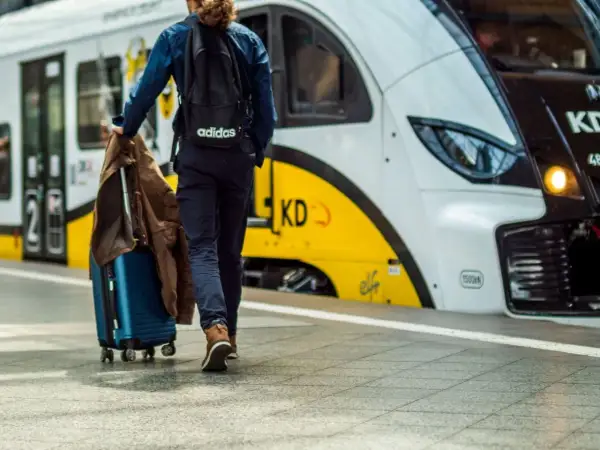 Podróżny z walizką i kurtką idzie w stronę pociągu KD. Rozkład PKP czeka na pasażerów.