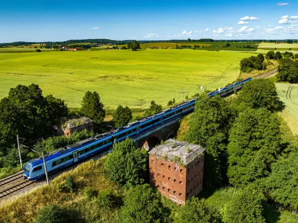 Szynobus Olsztyn Szymany mija ruiny fortu na tle zielonych pól i błękitnego nieba.