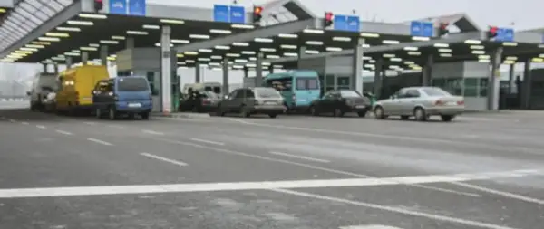 Kolejka samochodów na granicy polsko rosyjskiej. Widać budynki przejścia granicznego, sygnalizację świetlną i różne pojazdy.